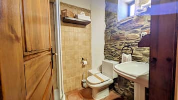 A bathroom with a stone wall, a wooden door, a toilet, a sink, and a shelf with towels.