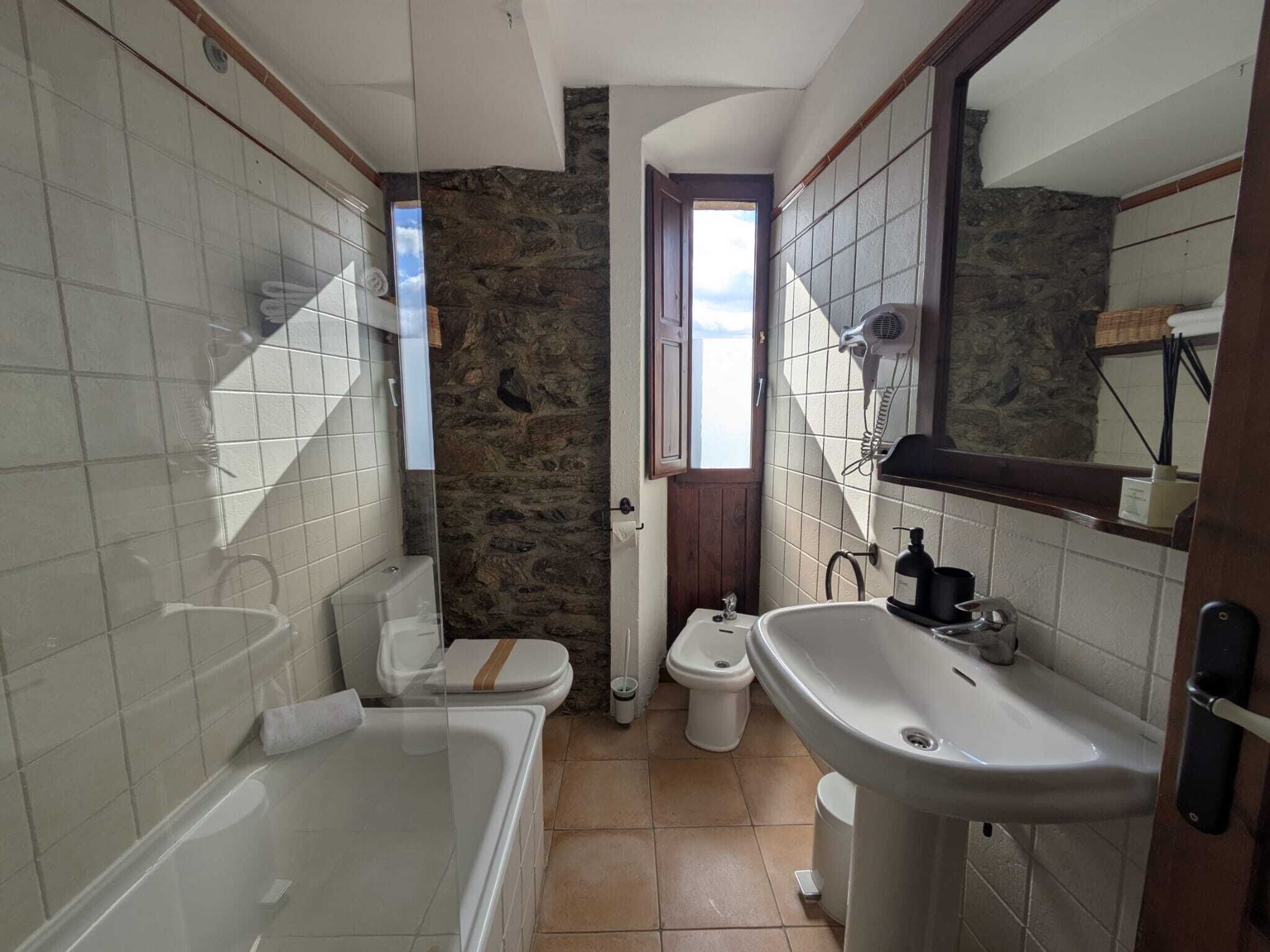 Un baño con bañera, inodoro y lavamanos. Cuenta con una pared de piedra y una ventana.