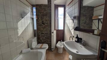 Un baño con bañera, inodoro y lavamanos. Cuenta con una pared de piedra y una ventana.