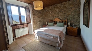 Un dormitorio con pared de piedra, techo de madera, cama con cabecera, mesita de noche, ventana con cortinas y radiador.