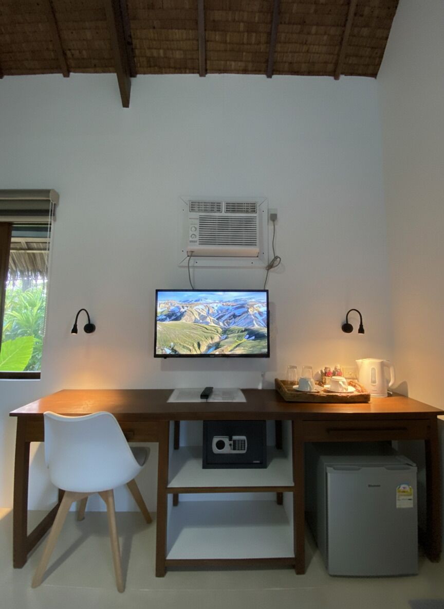Una habitación con un escritorio de madera, una silla blanca, un televisor, un aire acondicionado y un refrigerador pequeño.
