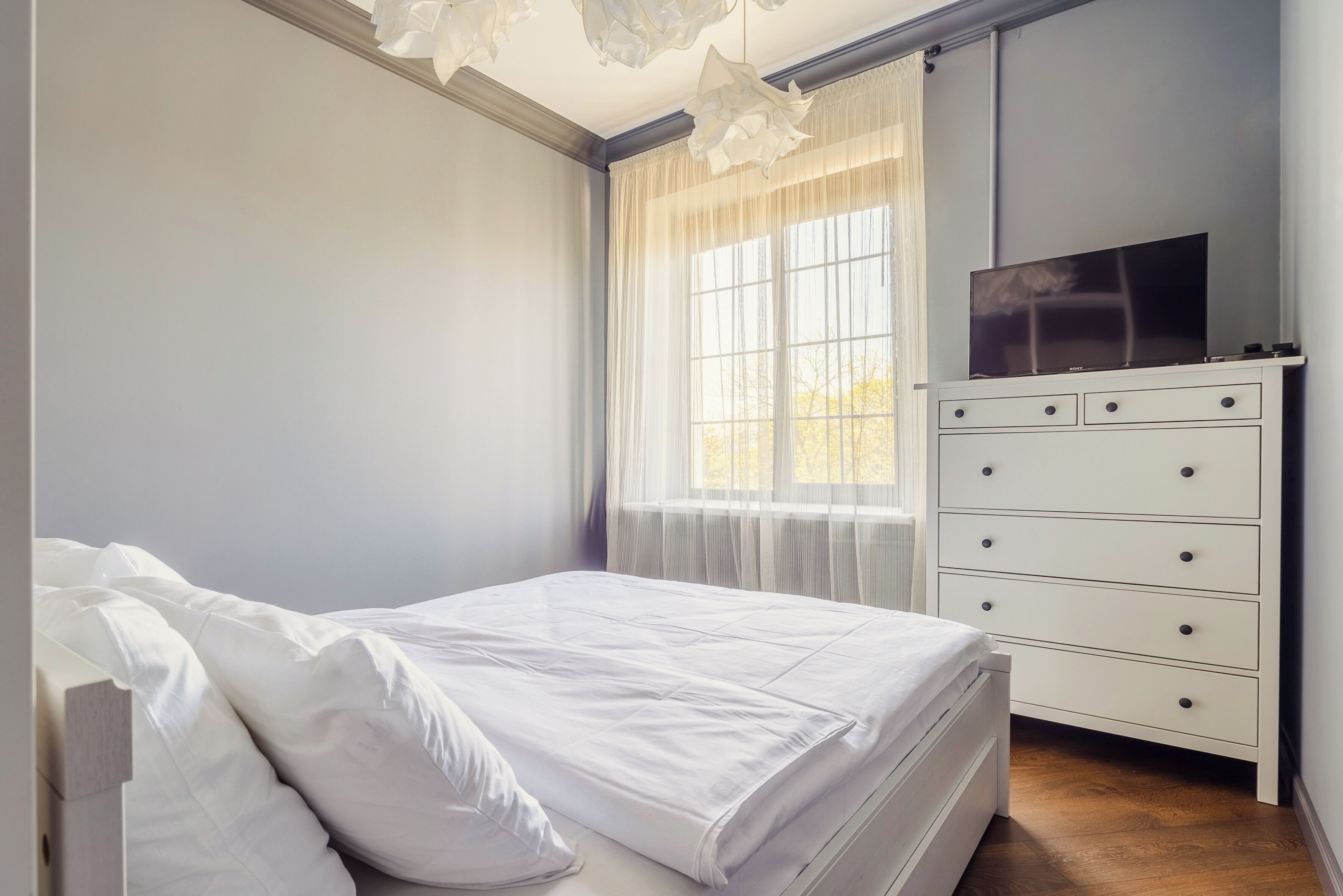 Une chambre à coucher avec un lit, une commode, un téléviseur posé sur le dessus, et une fenêtre avec des rideaux légers.