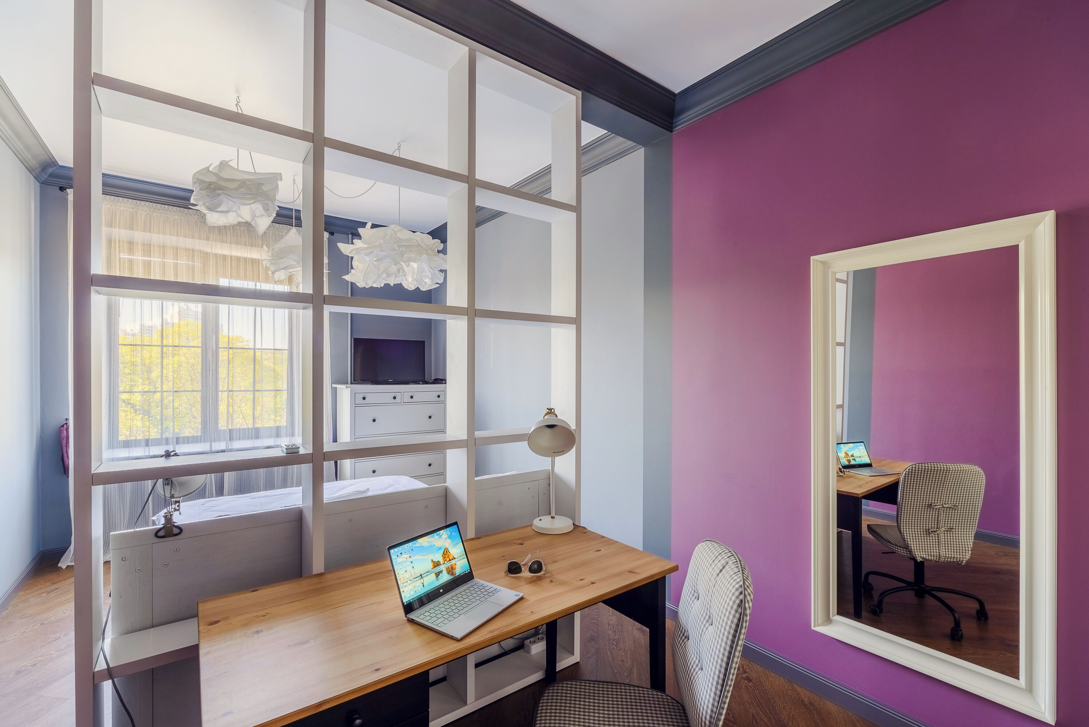 Une pièce avec un bureau en bois, un ordinateur portable, une paire de lunettes, une lampe, une chaise et un grand miroir sur un mur violet.