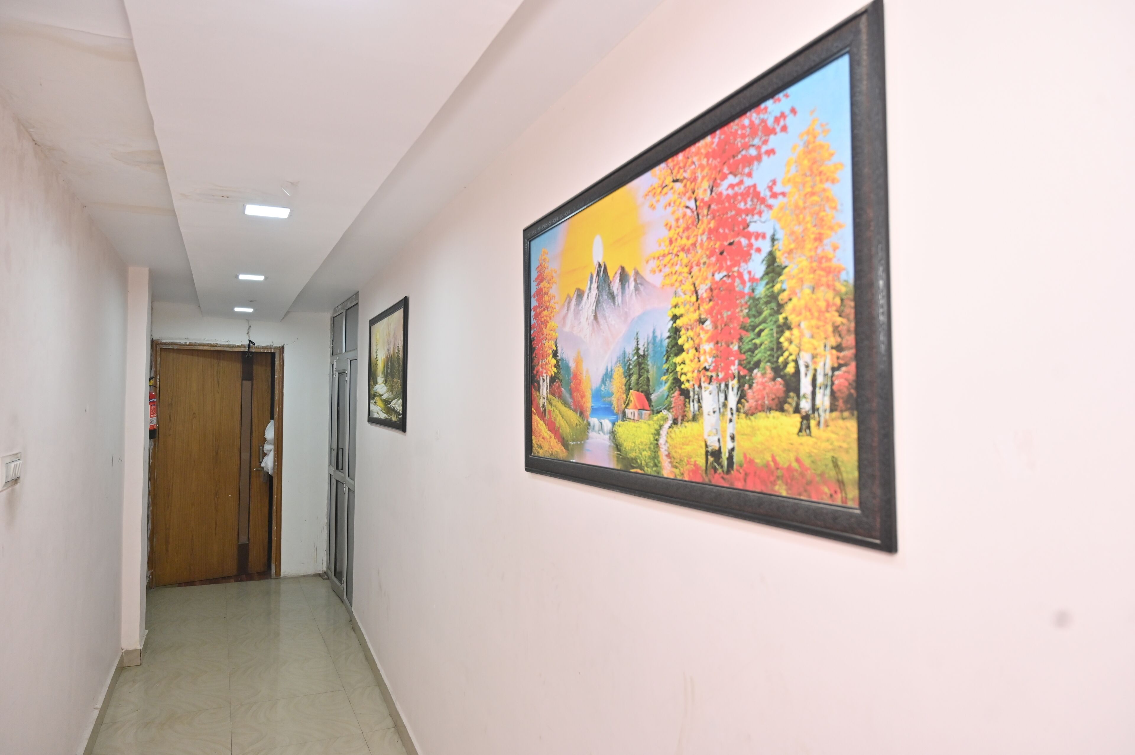 Un couloir avec une porte en bois, un tableau encadré représentant des arbres d’automne et un sol carrelé.