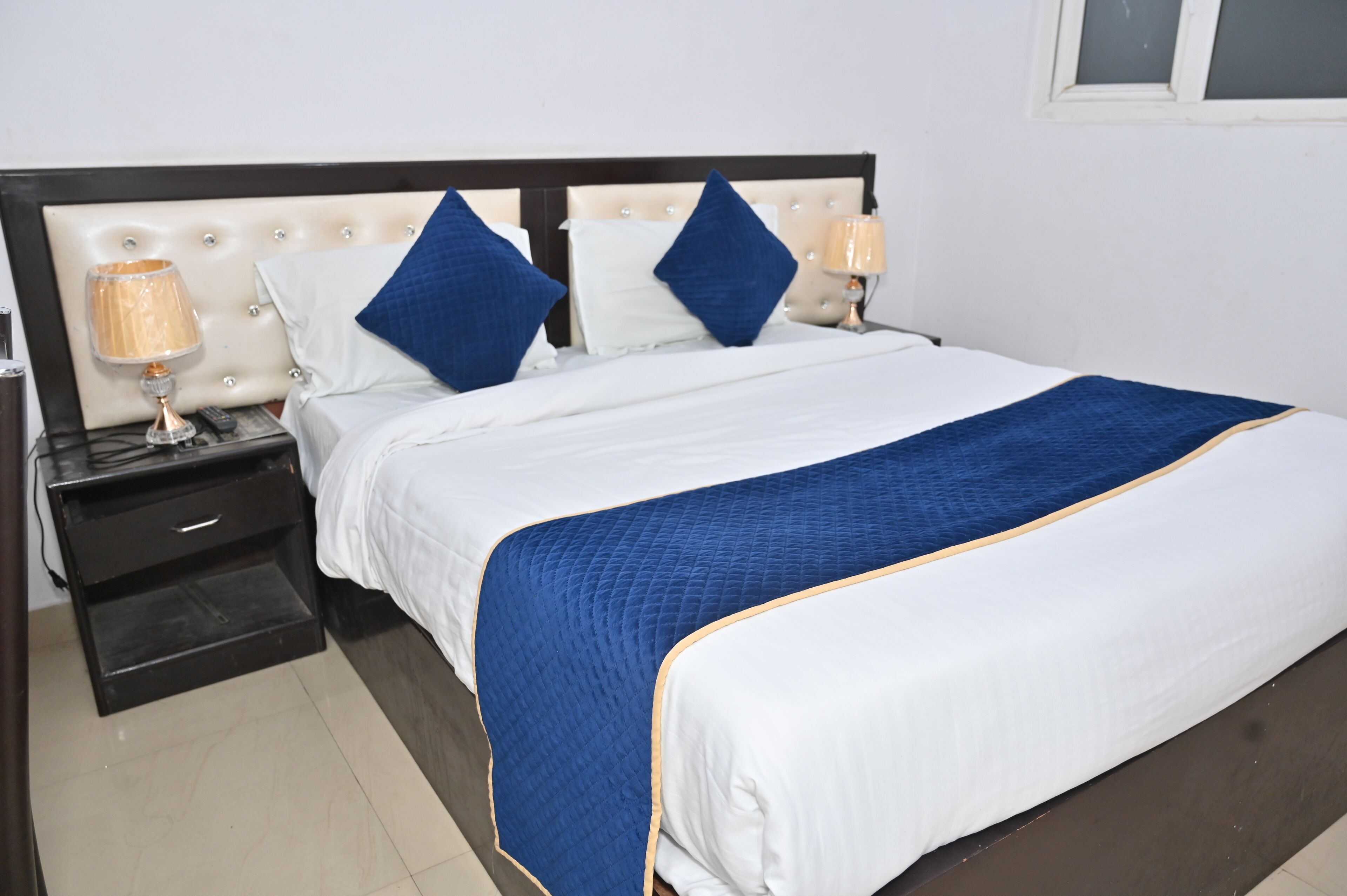 Una cama bien hecha con colcha azul y ropa de cama blanca, una mesita de noche de madera con una lámpara y una ventana sobre la cama.
