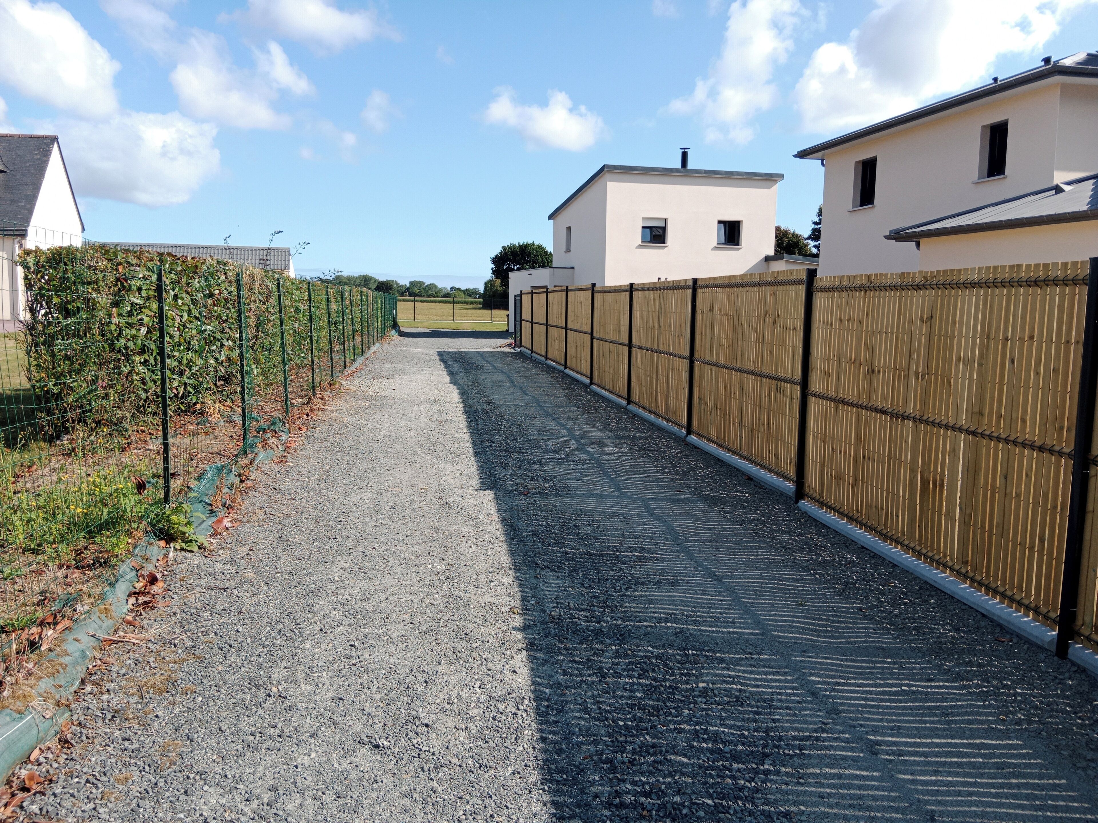 Un chemin de gravier mène à un bâtiment, avec une clôture en bois d’un côté et une clôture en métal de l’autre.