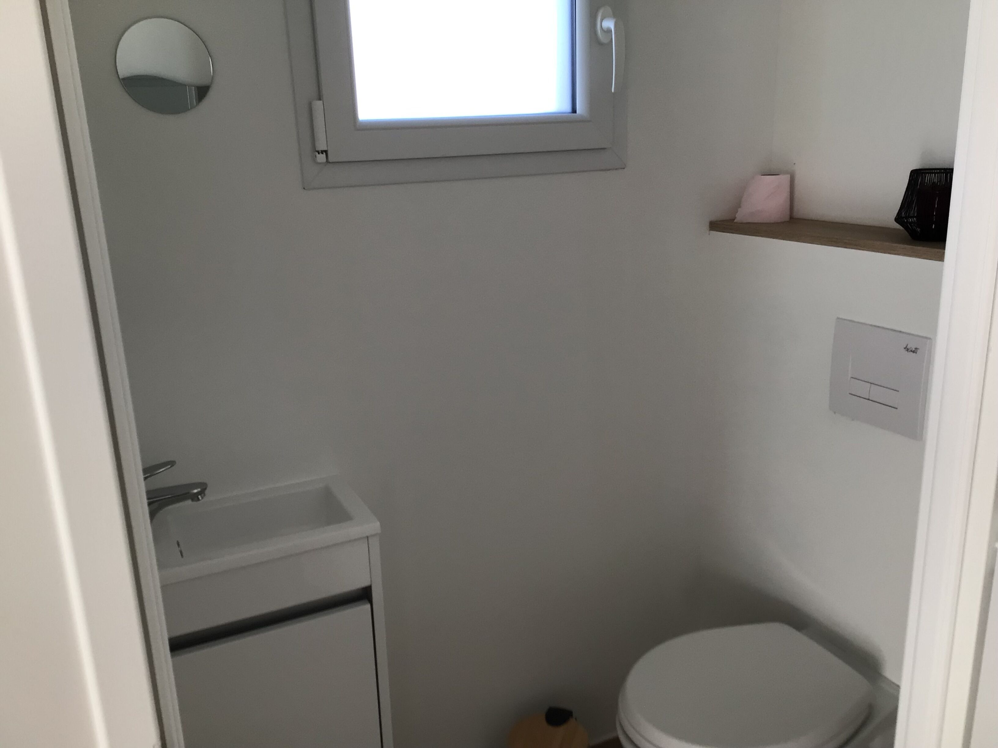 Un baño pequeño y limpio con inodoro y lavamanos blancos, y un espejo sobre el lavamanos. Hay una ventana sobre el inodoro y una repisa con un dispensador de pañuelos y un recipiente negro.