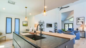 Una cocina moderna con una isla central, un comedor con sillas azules y una sala de estar con sofá y televisor.