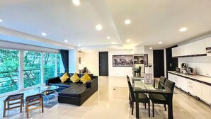 Een moderne woonkamer met een bank, eettafel en stoelen, een keuken met kasten, en grote ramen met uitzicht op bomen.
