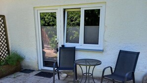 Eine Außenpatio mit zwei Stühlen und einem kleinen Tisch, einer Glasschiebetür und einer weißen Wand.