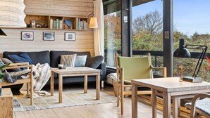 Interior acogedor de una cabaña con techo de madera, un sofá, una mesa y una silla. Se aprecia una estantería con libros y marcos con fotografías.