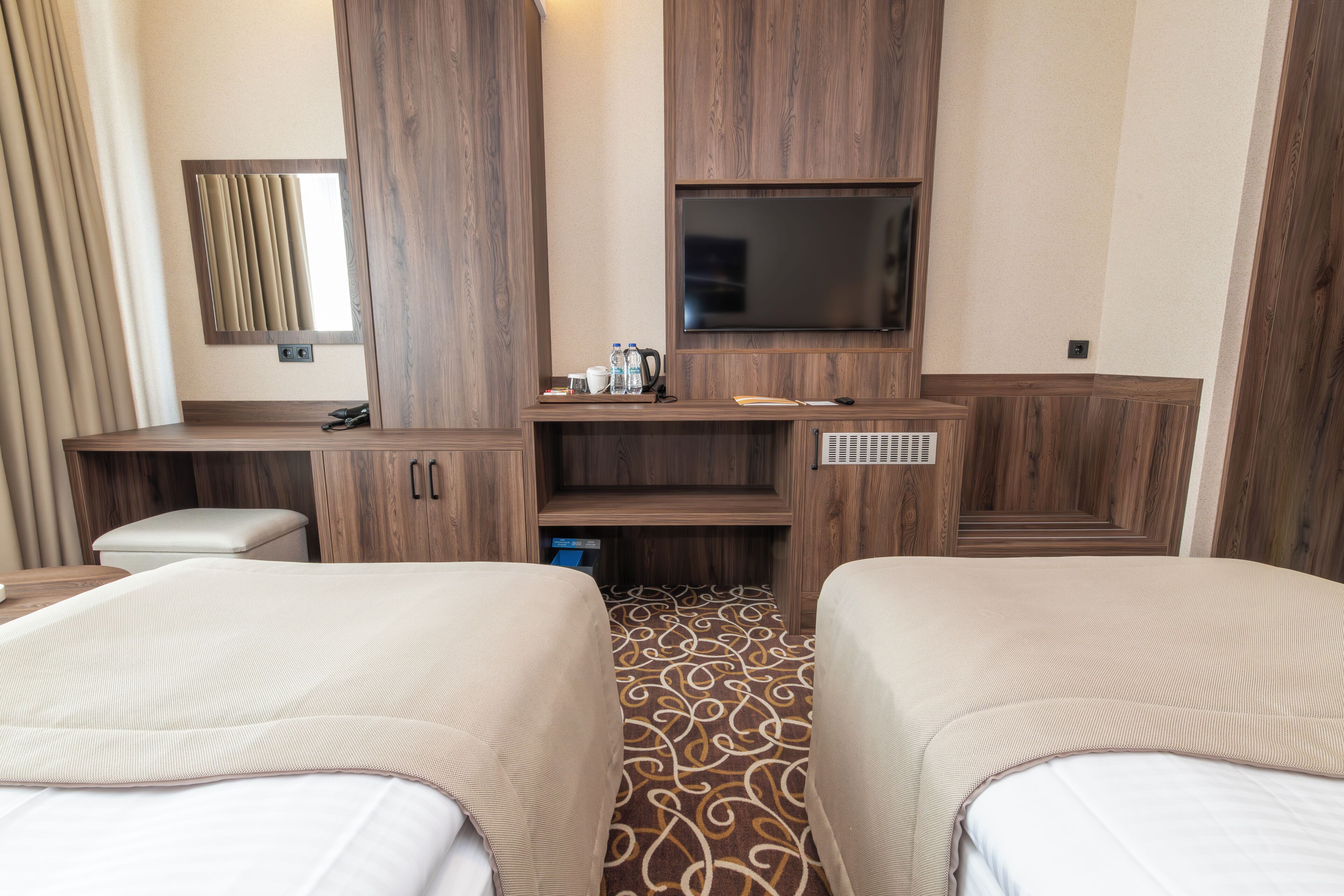 Habitación de hotel con dos camas, un televisor montado en la pared, un escritorio con silla y una pequeña zona de kitchenette.