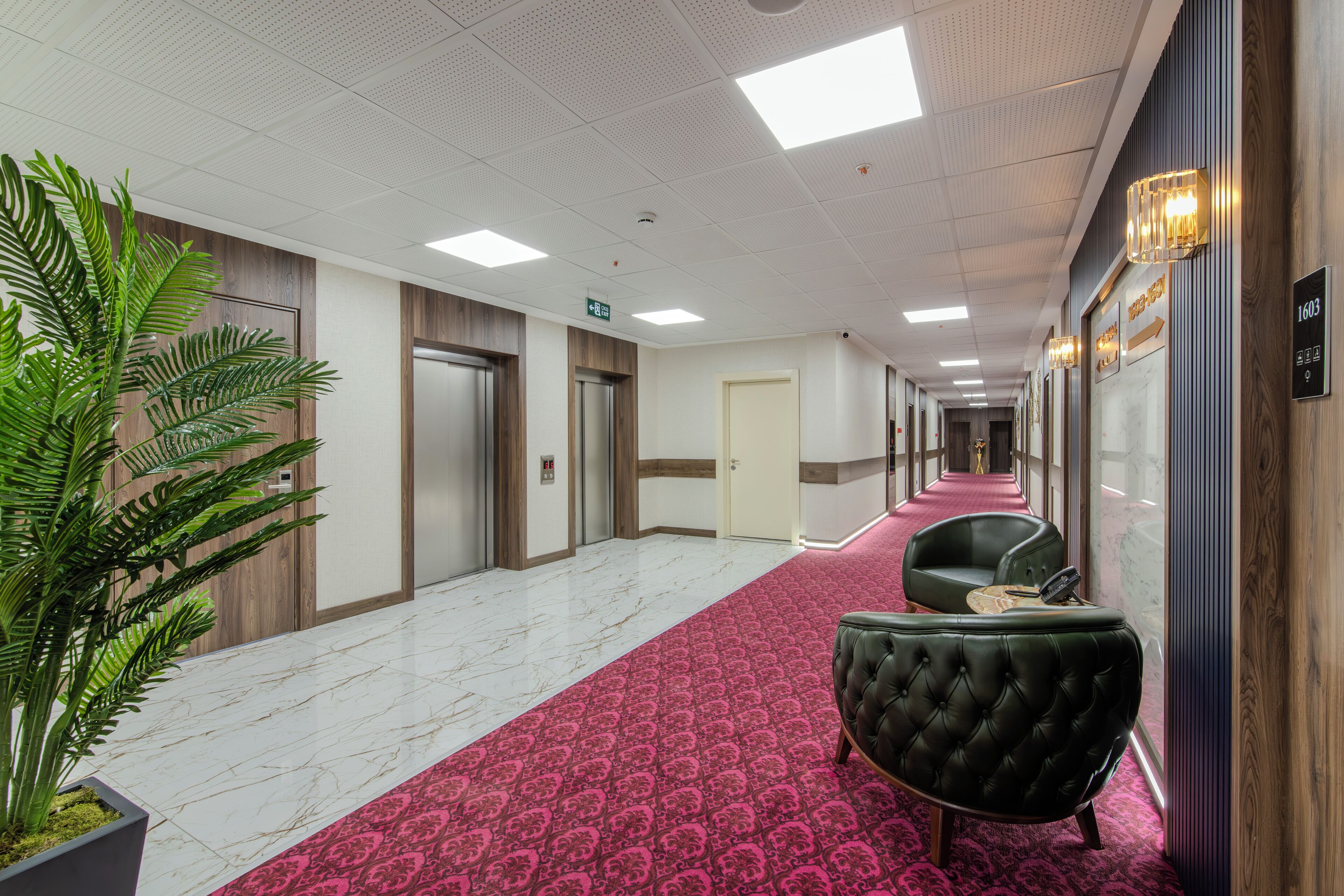 Een hotelgang met een rood tapijt, een zwarte leren fauteuil en een potplant.