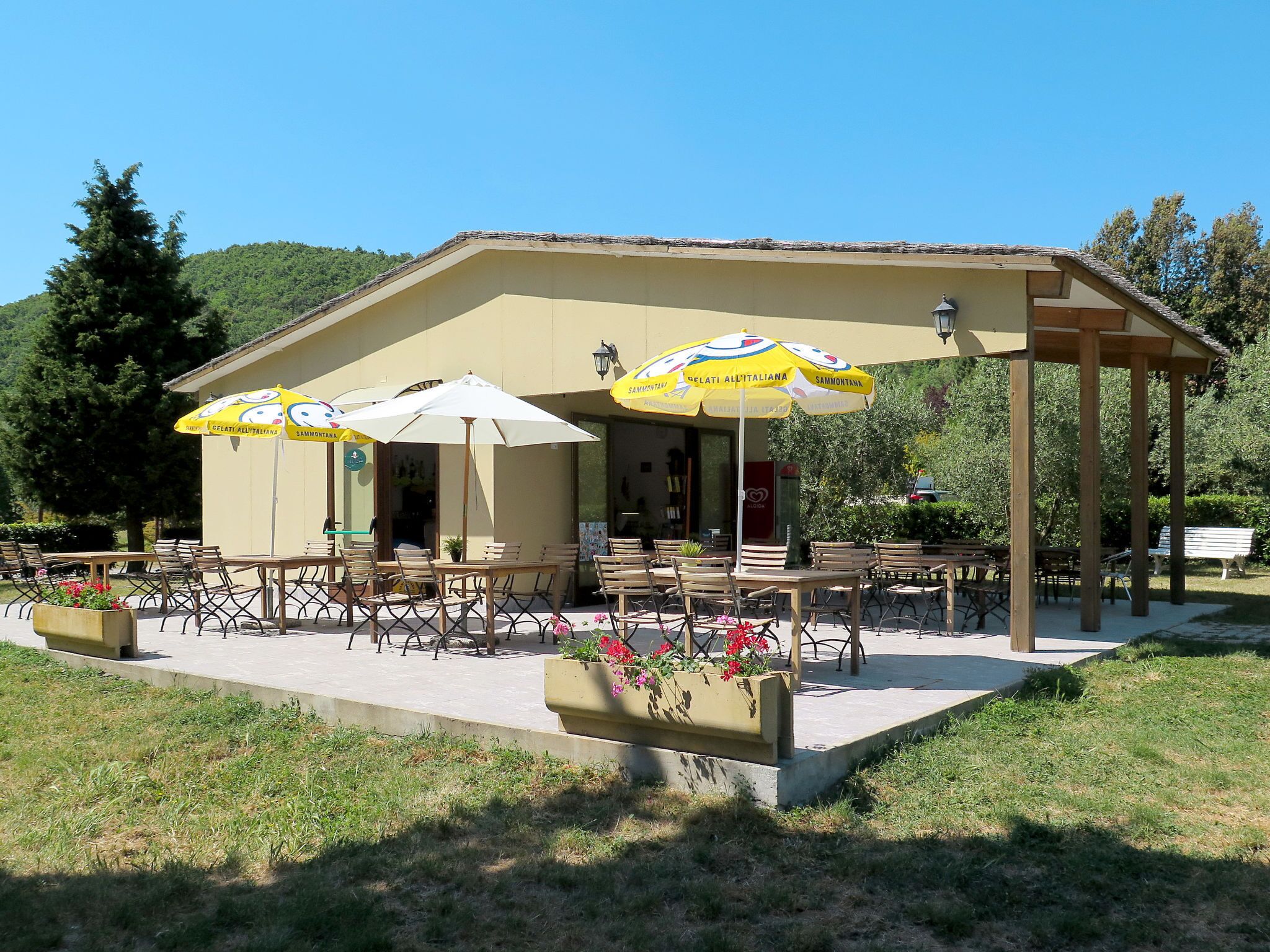 A building with outdoor seating and umbrellas, surrounded by greenery and a clear sky.