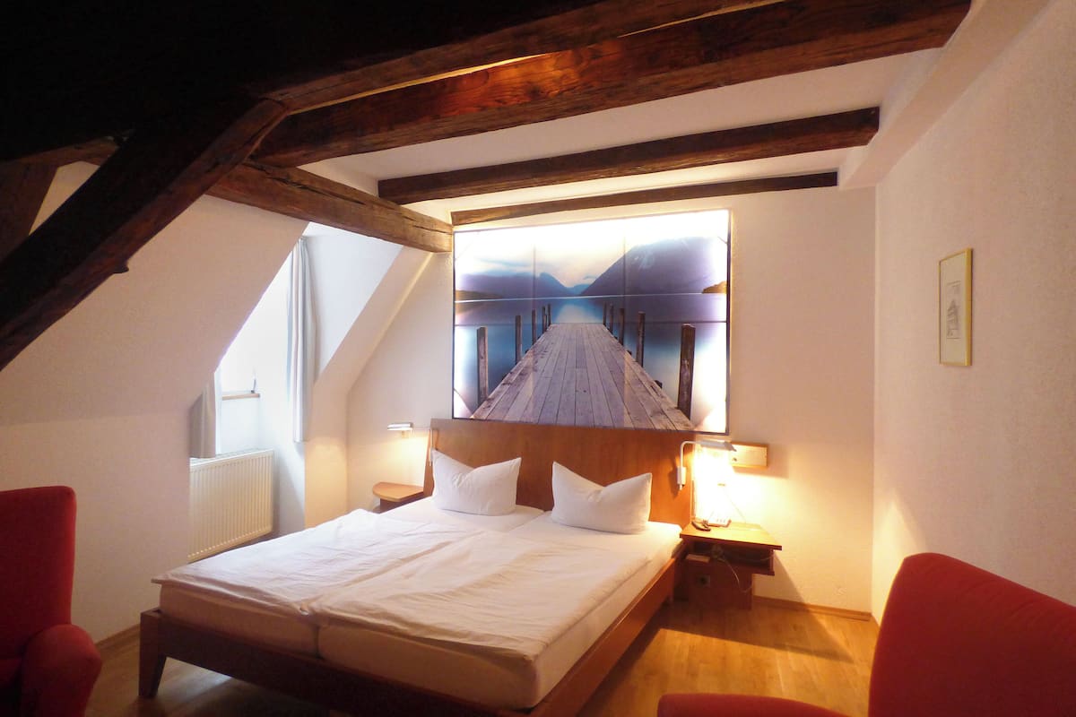 A bedroom with a wooden ceiling, a bed with white bedding, two bedside tables with lamps, and a window with a view of a mountain.