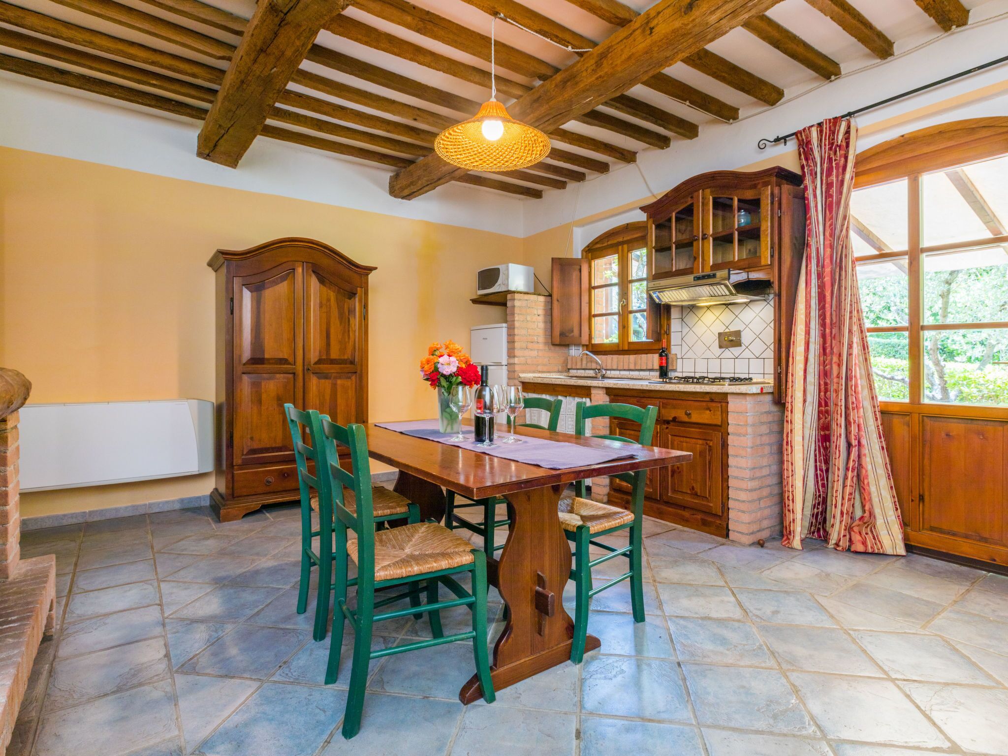 A dining area with a wooden table and chairs, a vase with flowers, and a kitchen in the background.