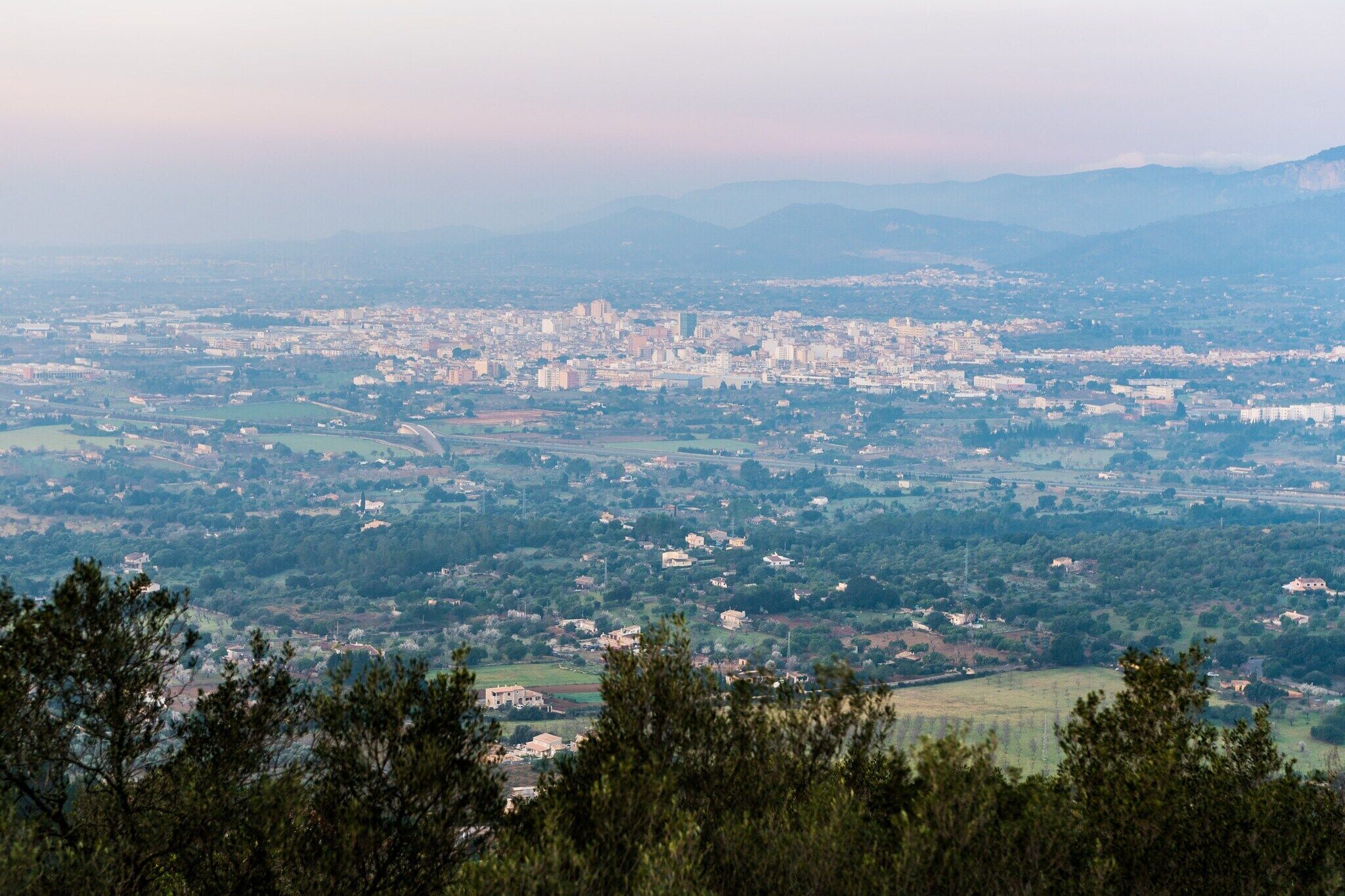 Vista panorámica de una ciudad con un centro urbano denso, rodeada de campos verdes y montañas al fondo.