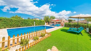 A swimming pool with a wooden fence, a grassy lawn, and a building with a red umbrella in the background. - Can Tomeu - Villa With Private Pool In Inca (Inca, Illes Balears)