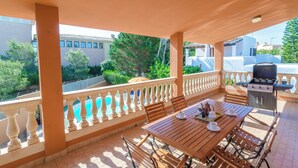 An outdoor patio with a wooden table and chairs, a grill, and a view of a swimming pool and buildings.