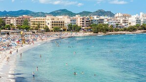 A beach with people swimming and sunbathing, a promenade with buildings, and a mountainous backdrop.