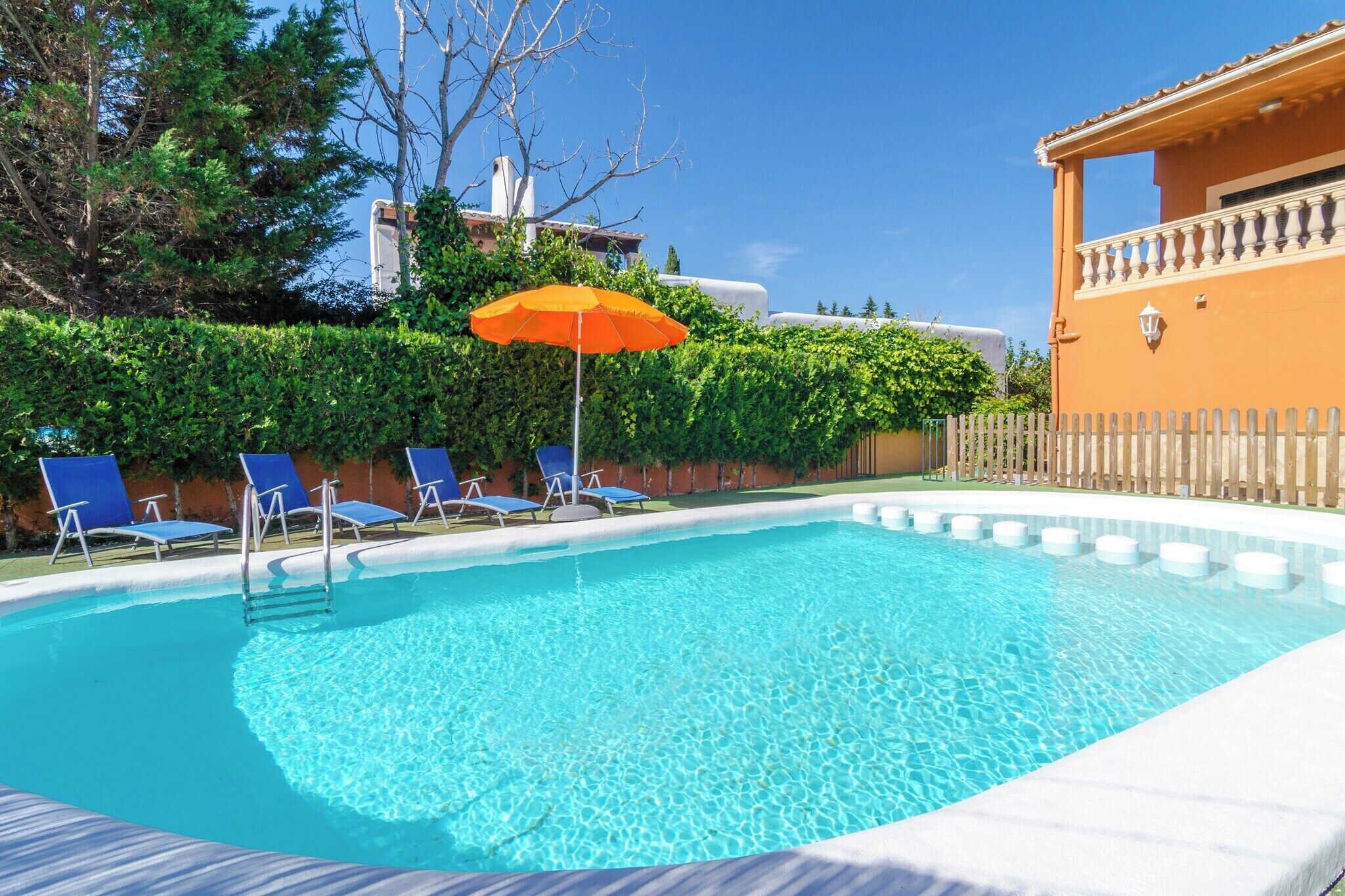 A clear swimming pool with blue water, surrounded by a wooden fence and a building with a tiled roof.