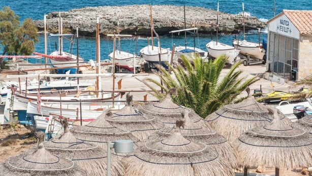 A coastal marina with boats docked, a building labeled 'NAUTICA DES ILLOT', and thatched umbrellas.