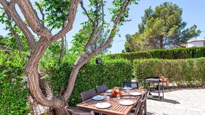 An outdoor dining area with a wooden table and chairs, surrounded by greenery and a hedge.