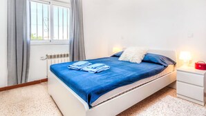 A bedroom with a bed, a window with curtains, a radiator, a nightstand with a lamp, and a red bag.