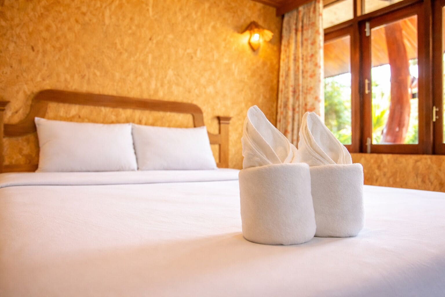 A hotel room with a neatly made bed, a wall-mounted light, and a window with curtains.