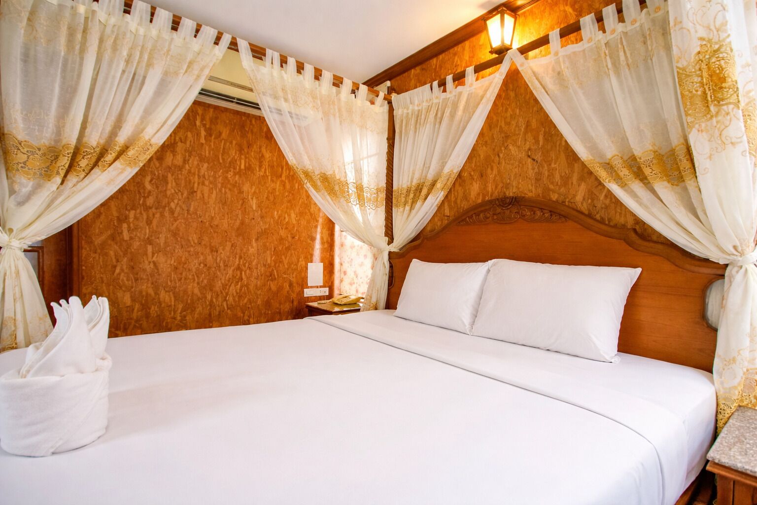 Habitación de hotel con cabecera de madera, ropa de cama blanca y cortinas de gasa con diseños dorados.