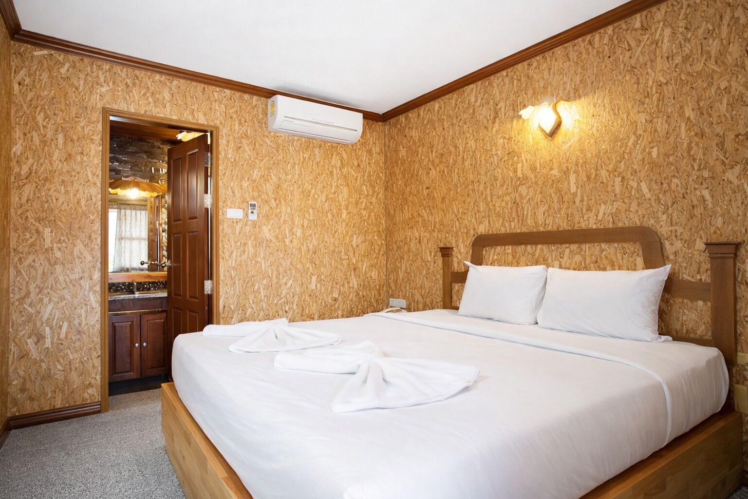 A hotel room with a wooden wall, a bed with white linens, a wooden headboard, and an air conditioning unit.