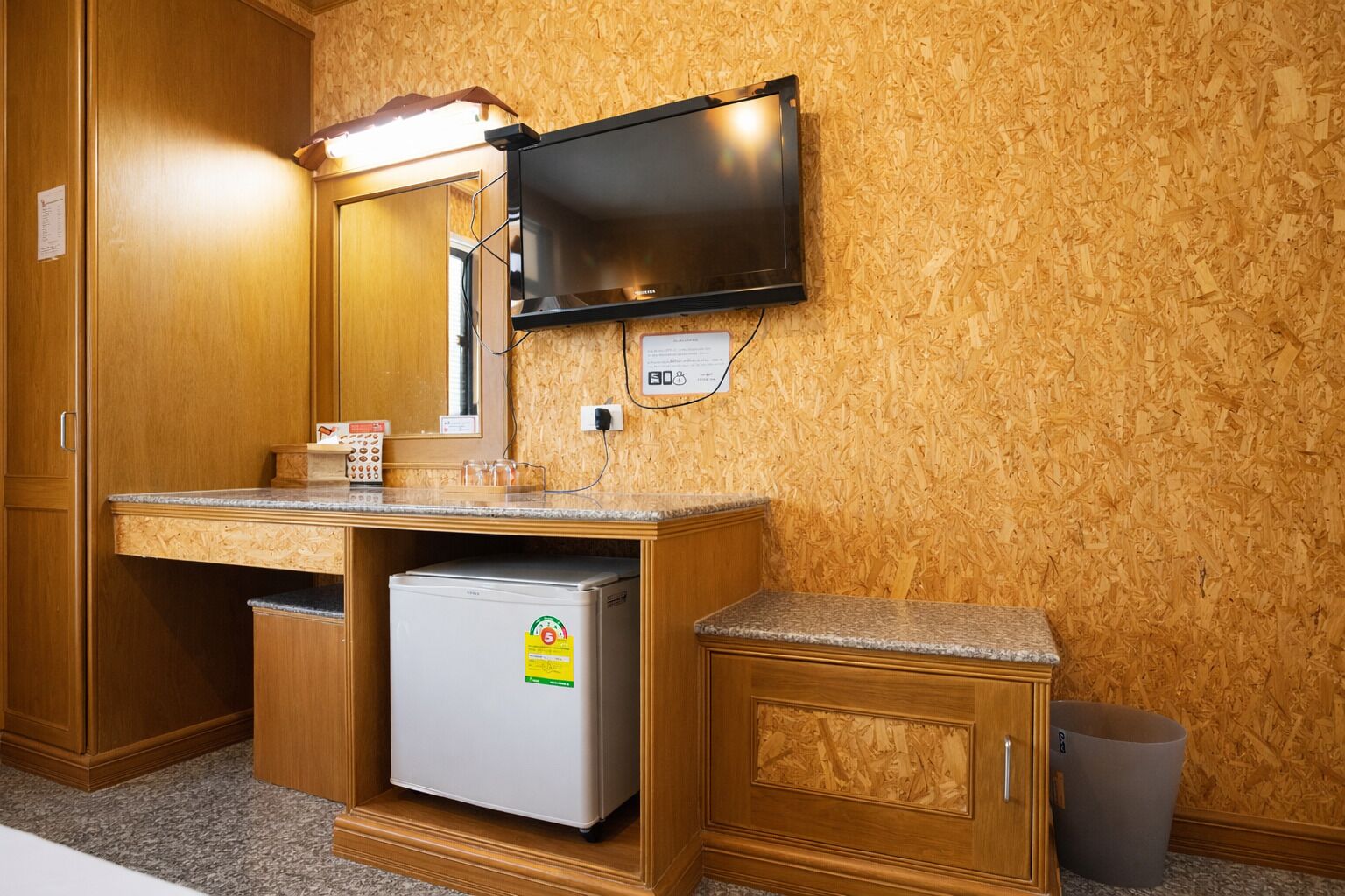 A compact bathroom with a wooden vanity, a small refrigerator, a mirror, and a television mounted on the wall.