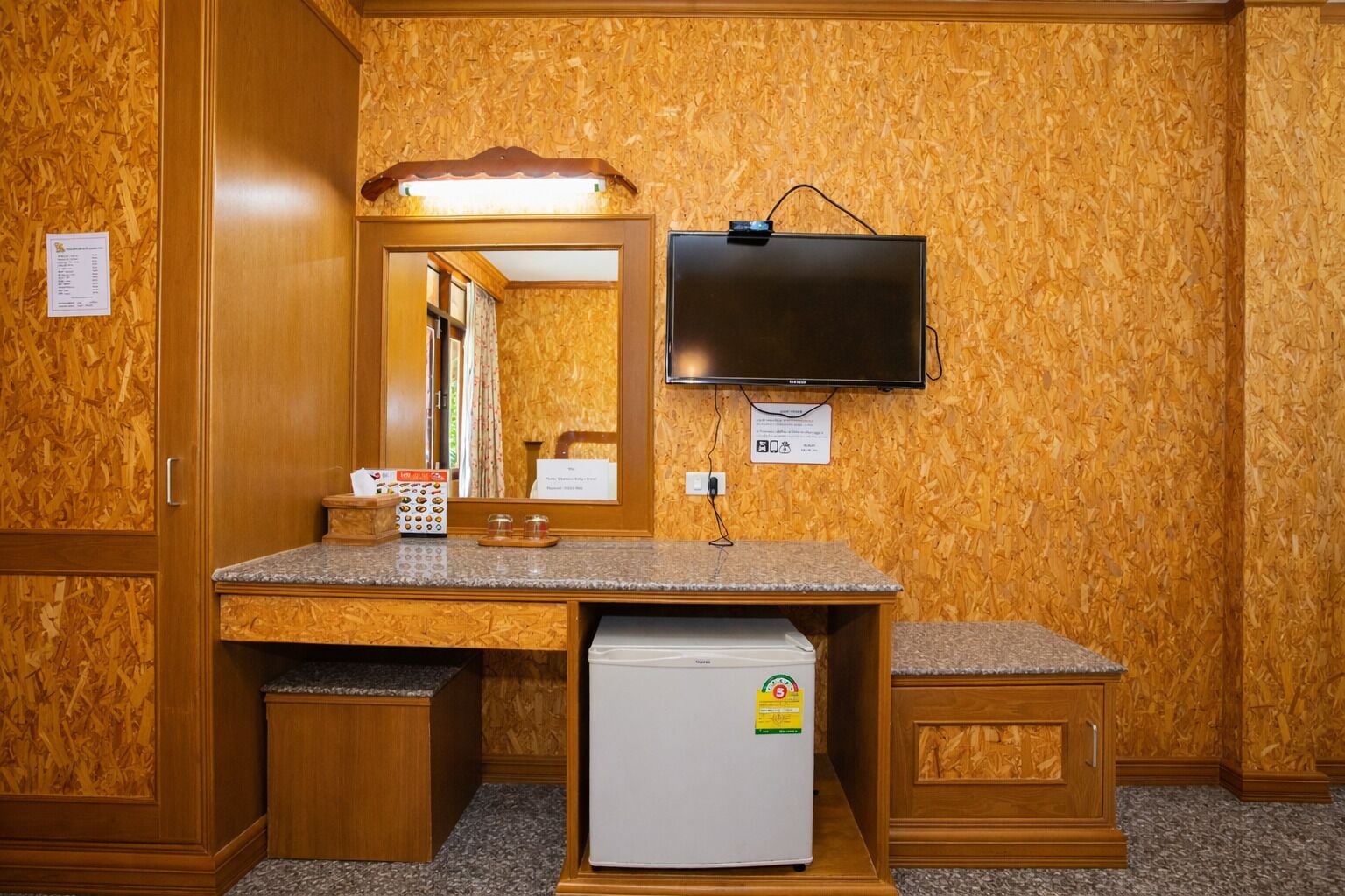 A hotel room with a wooden wall, a flat-screen TV mounted above a counter, a refrigerator, and a mirror with a light above it.