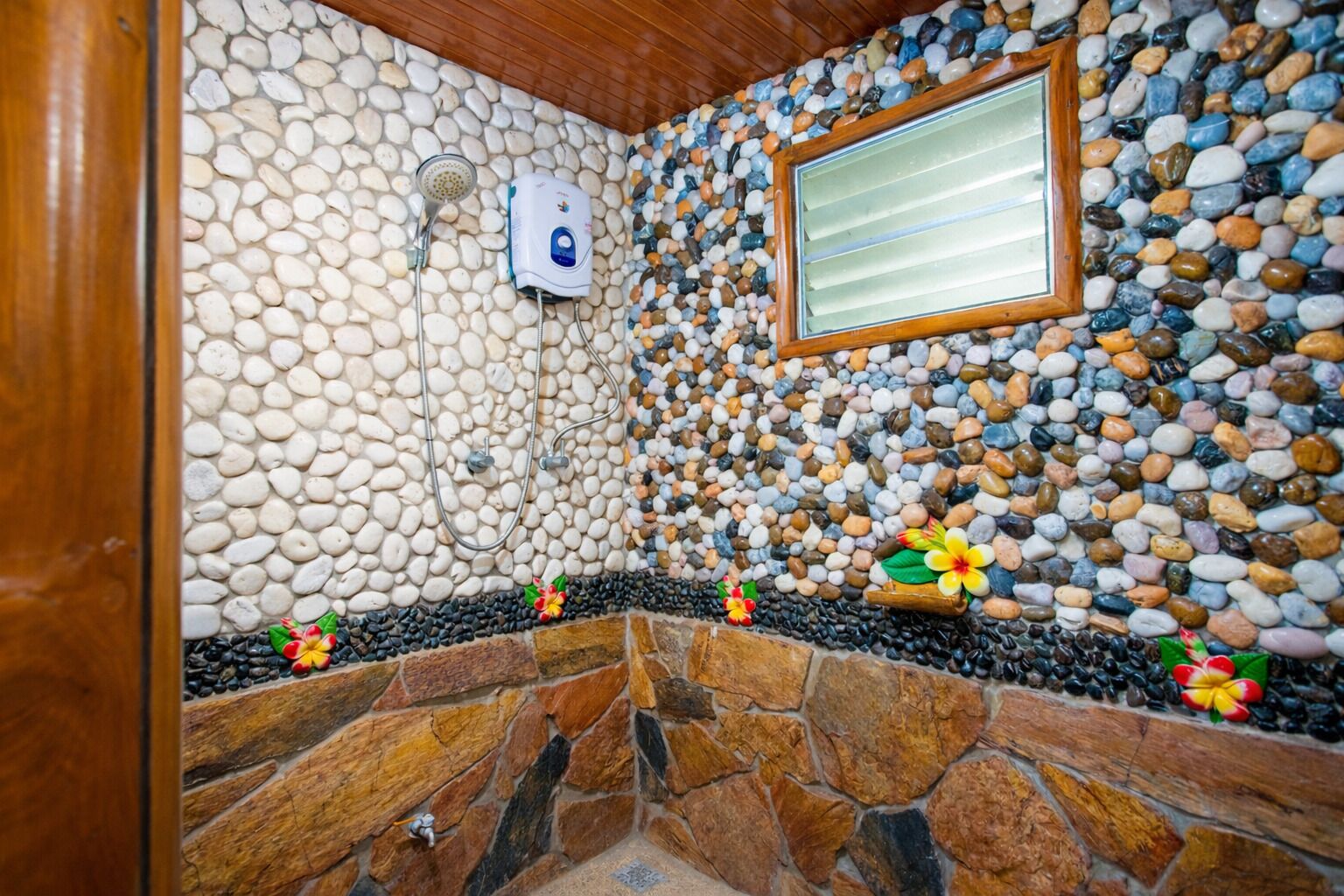 A stone-walled bathroom with a shower, a wooden-framed window, and decorative flowers.