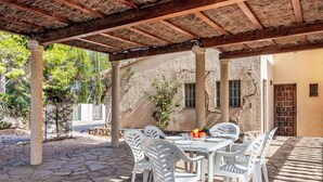 An outdoor patio with stone flooring, white plastic chairs, and a table set with fruit and glasses.