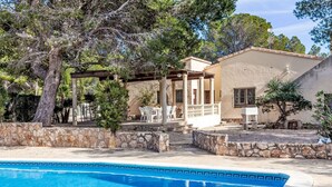 A swimming pool with a stone border, a pergola with outdoor seating, and a building with a tiled roof.