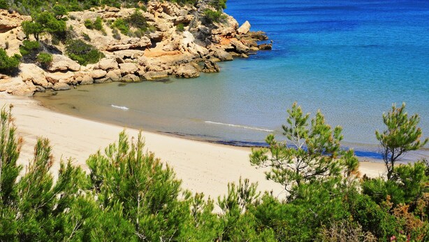 A coastal landscape with a rocky shoreline, clear blue sea, and a sandy beach.