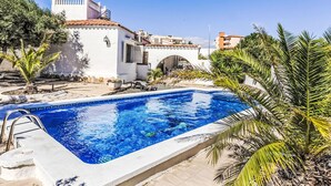 A swimming pool with a clear blue water, surrounded by palm trees and a white building with a red-tiled roof. - Vacation home Arcada by Interhome (Miami Platja)