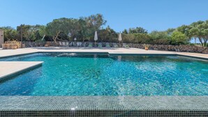 Une piscine d’un bleu éclatant, bordée de carreaux, entourée de chaises longues et de parasols, le tout se dressant sur un fond de verdure composé d’arbres et d’un mur de pierre.