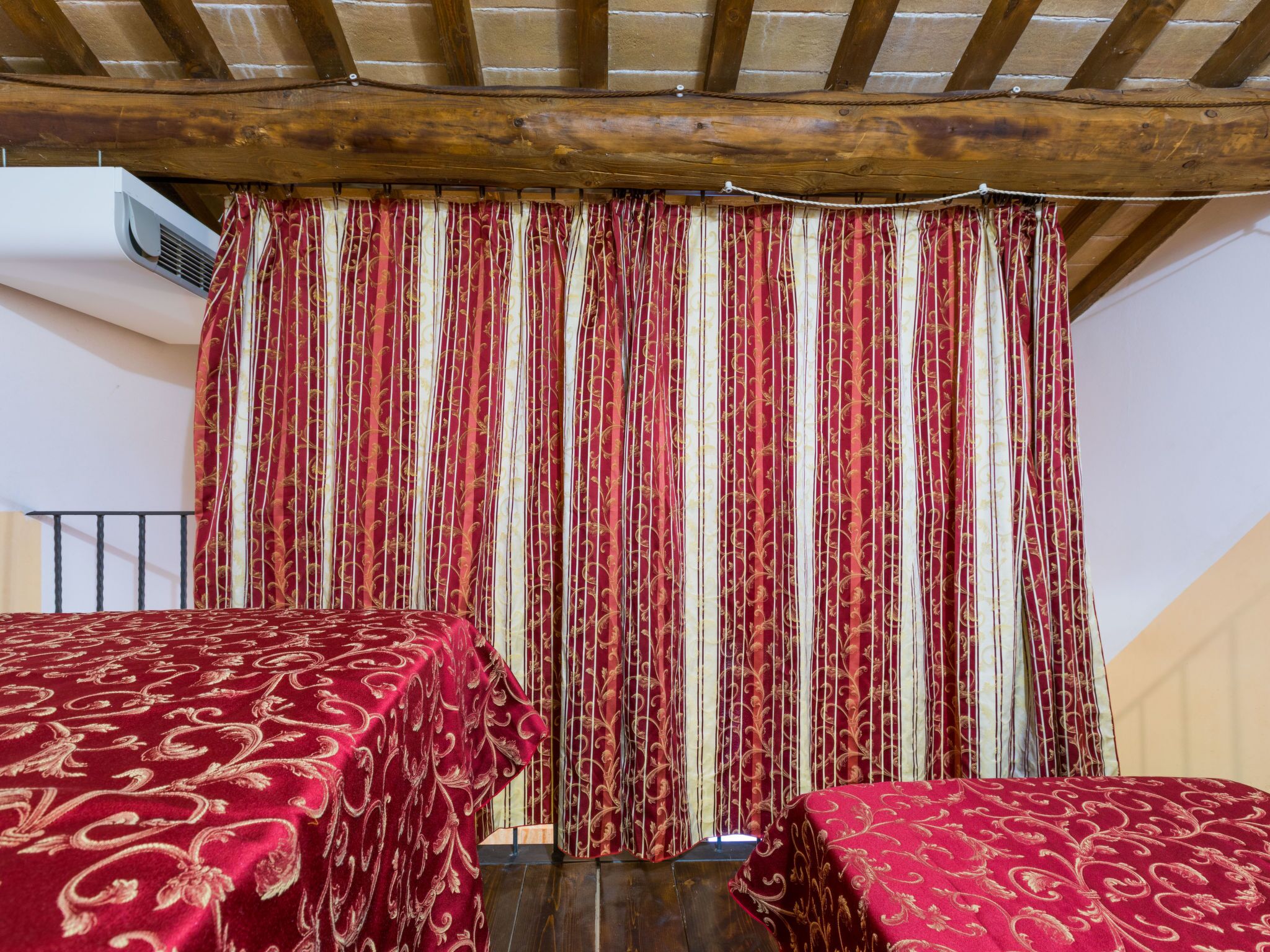 A room with two beds covered in red patterned bedding, a wooden ceiling, and a striped curtain.