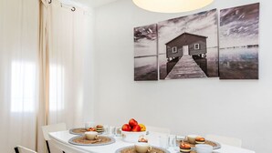 A dining area with a white table set for four, surrounded by white chairs. There are three paintings on the wall depicting a house by the water.