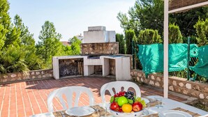 An outdoor patio with a stone barbecue, white plastic chairs, and a table set for two with a bowl of fruit.