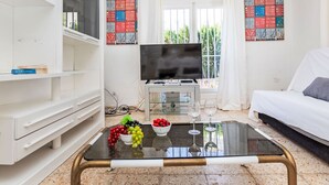 A modern living room with a flat-screen TV, a glass coffee table with fruit and wine glasses, a white sofa, and a TV stand with a small TV.