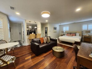 A modern living room with a leather sofa, a round coffee table, a dining area with a table and chairs, a bedroom with a bed and nightstands, and a kitchen area. - Carriage House B at the Hamilton House (Savannah)