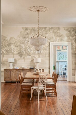 A dining area with a wooden table, white chairs, a chandelier, and a wall with a tree mural. - Hamilton House - 5 Bed Historic Downtown House w/ Courtyard & Parking (Savannah)
