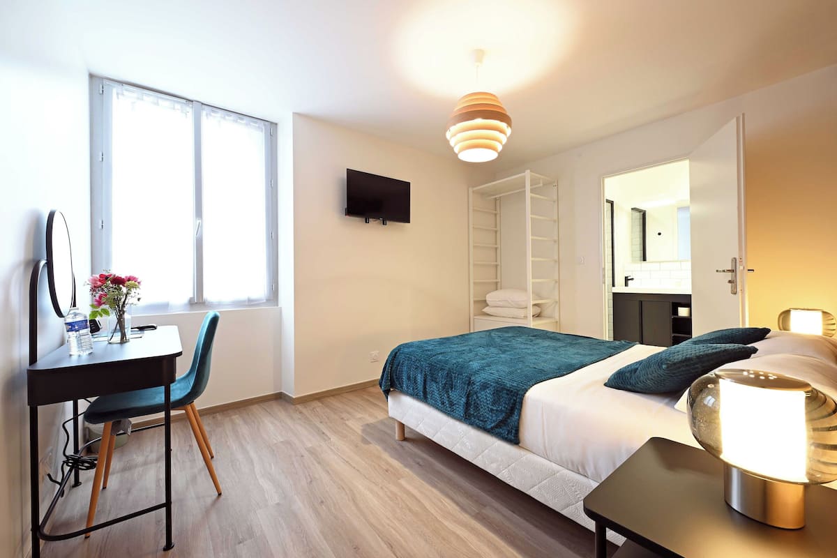 A modern hotel room with a bed, a desk with a chair, a TV, and a large window.