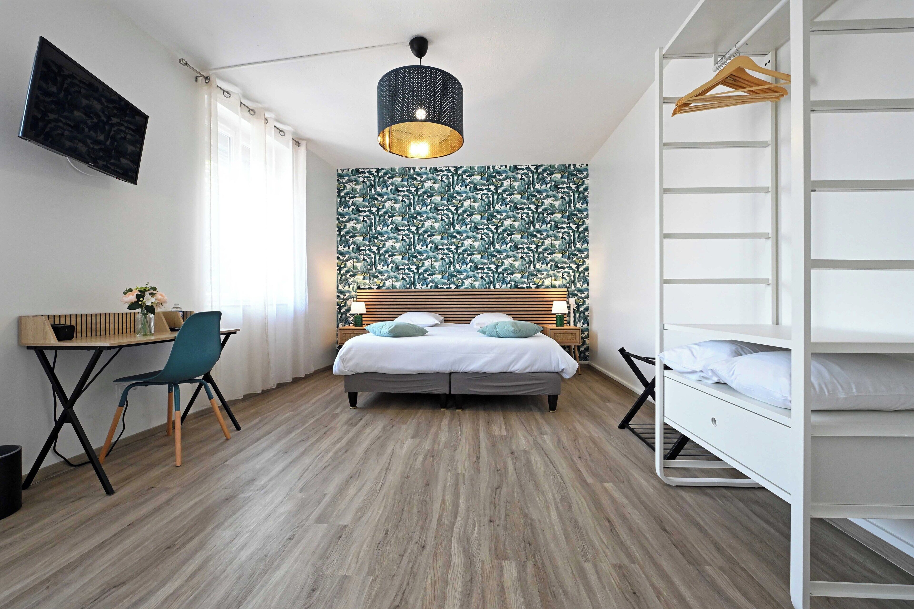 A modern bedroom with a bed, a desk, and a chair. There is a TV mounted on the wall and a hanging light fixture.