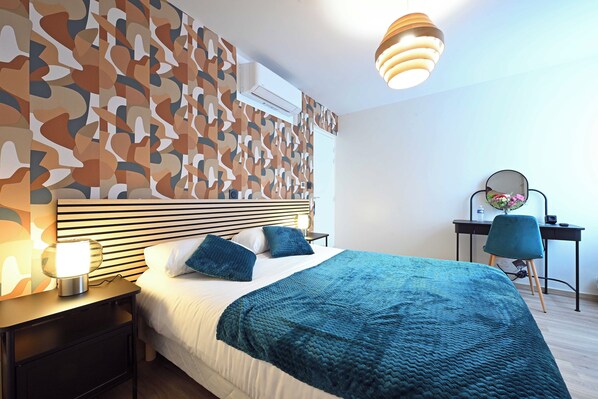 A modern hotel room with a patterned wall, a bed with a teal blanket, a bedside lamp, a desk with a chair, and a hanging light fixture. - Hôtel le Terminus et son restaurant Côte à Côte (Autun)