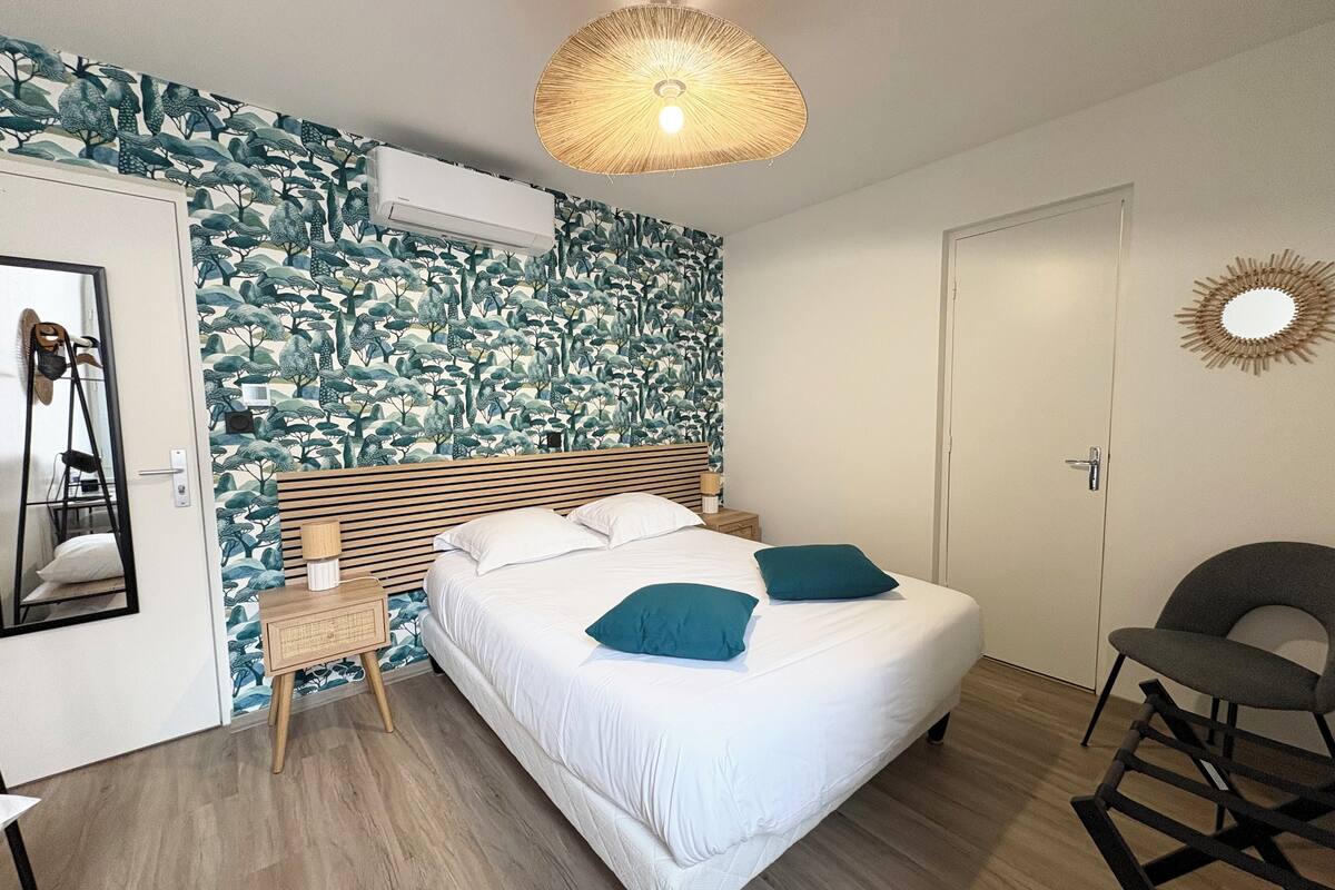 A bedroom with a bed, a bedside table, a chair, a wall-mounted air conditioner, and a decorative mirror.