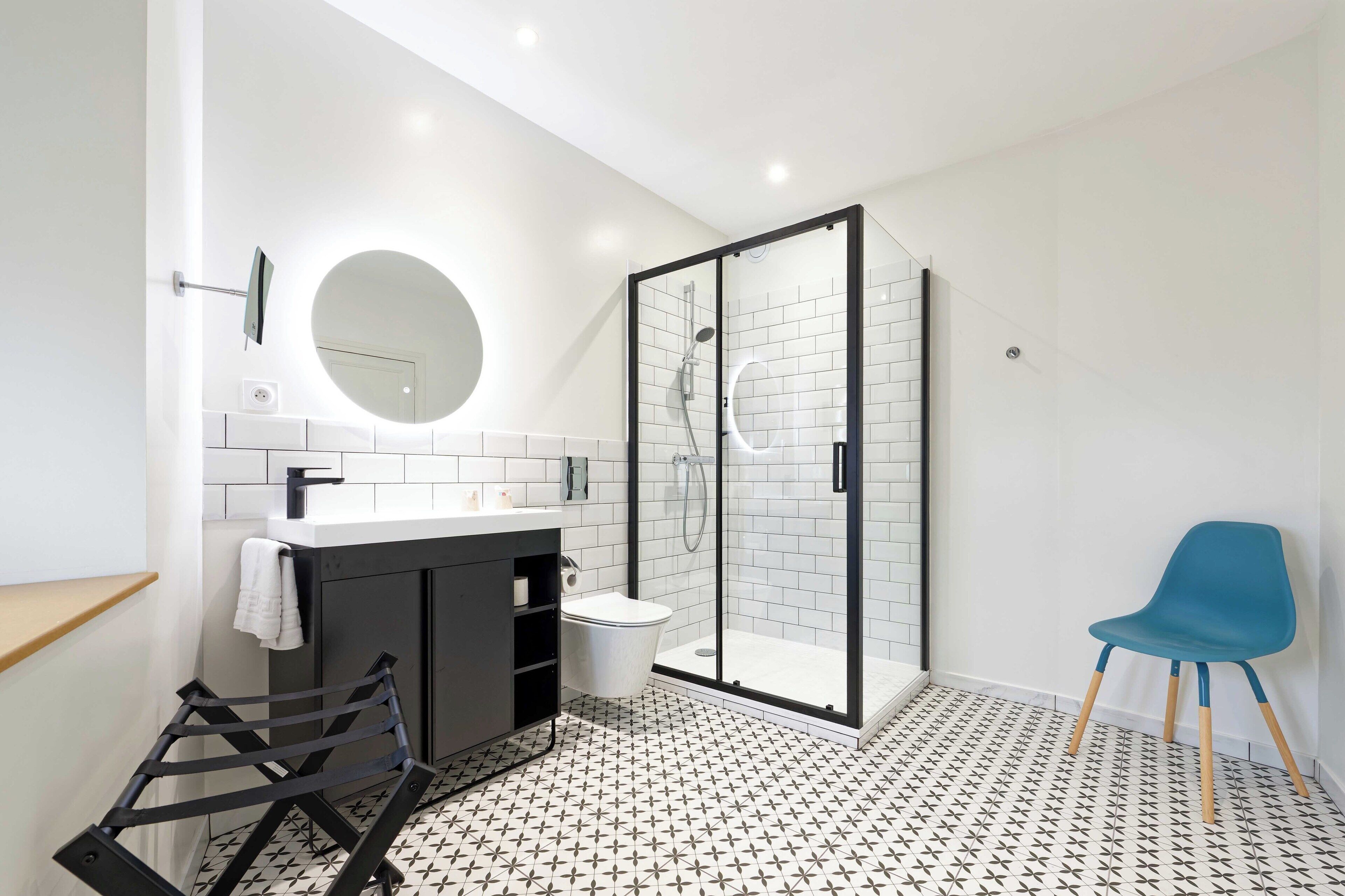 Un baño moderno con ducha envuelta en vidrio, espejo redondo, lavamanos blanco y silla azul.